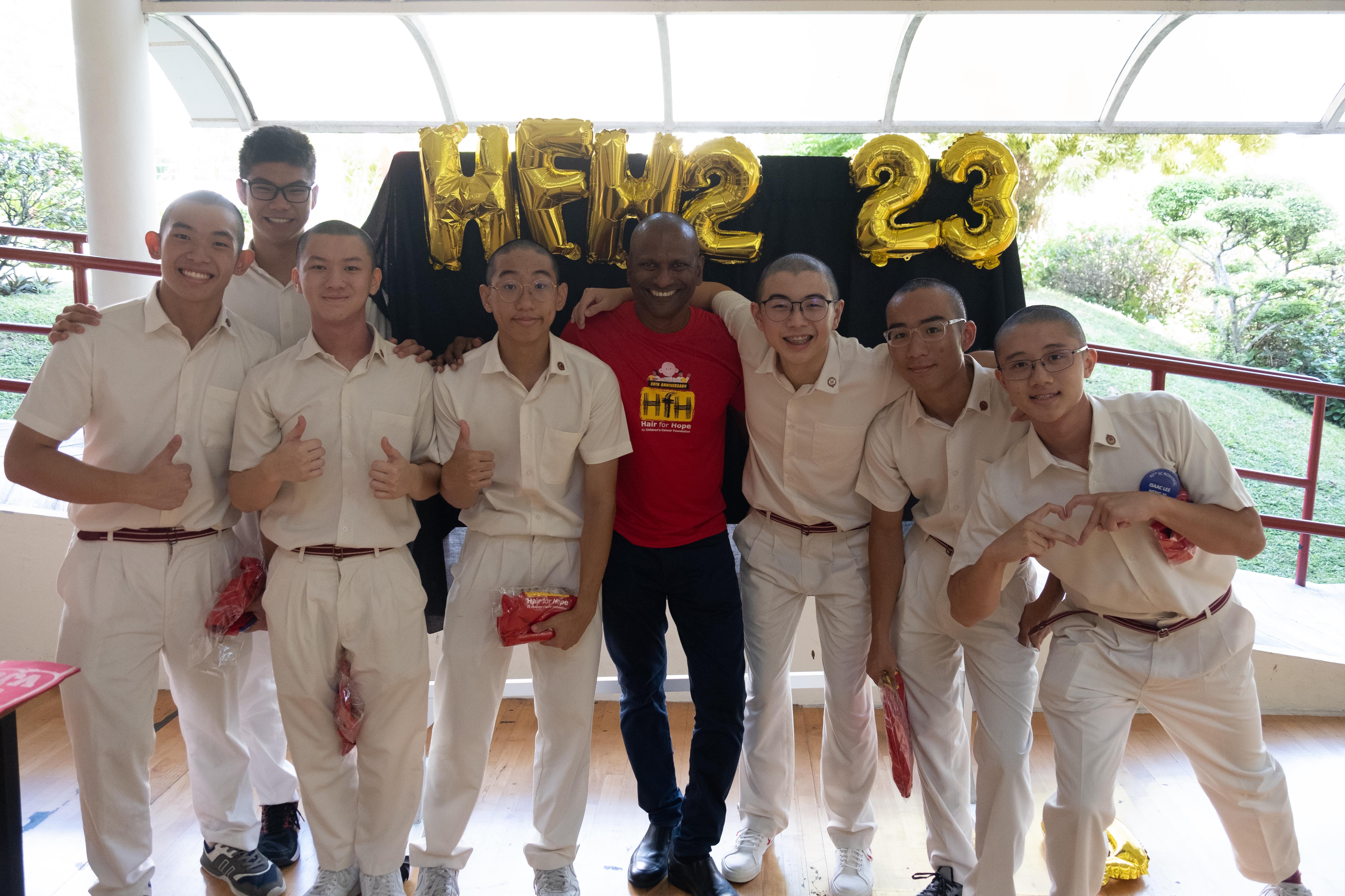 Group of shaved-head students in white uniforms with an adult in a red Hair for Hope shirt, posing before gold HFH 23 balloons.